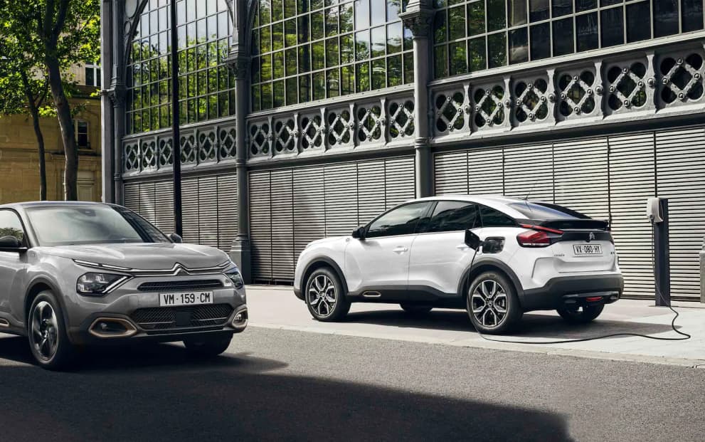 Front of a silver Citroen parked side by side with a white Citroën C4 plugged into a charging station