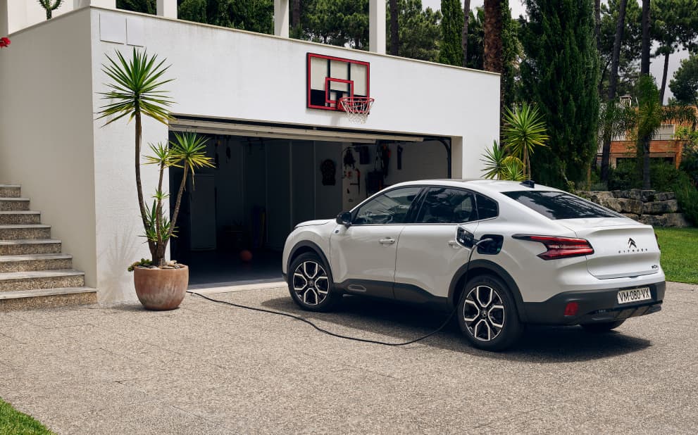 Angled side view of a white Citroen C4 X parked infront of a garage plugged into charge